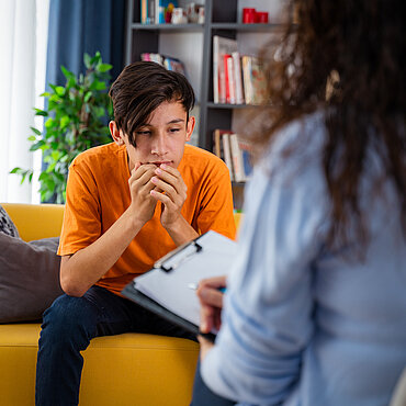 Ein Junge sitzt nachdenklich auf einem Sofa und spricht mit einer beratenden Person.