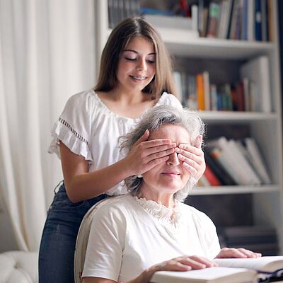 Ein Mädchen hält einer sitzenden Frau von hinten die Augen zu