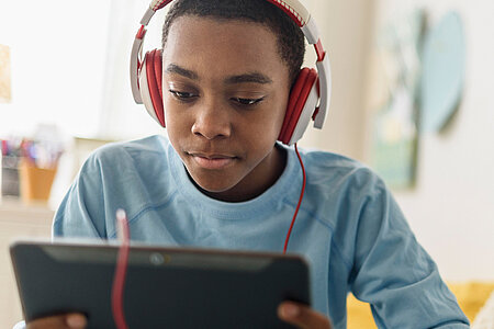 Jugendlicher sitzt entspannt auf einem Bett und schaut konzentriert auf ein Tablet, während er rote Kopfhörer trägt. 