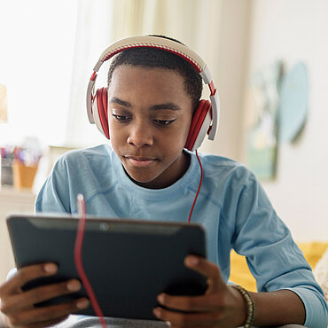 Jugendlicher sitzt entspannt auf einem Bett und schaut konzentriert auf ein Tablet, während er rote Kopfhörer trägt.