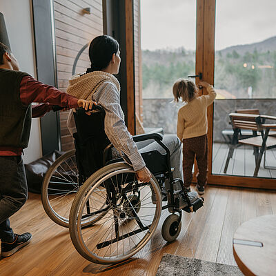 Zwei Kinder und eine Frau im Rollstuhl zusammen in einer Wohnung, ein Kind öffnet die Balkontür, das zweite schiebt den Rollstuhl