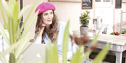Eine Frau mit Hut sitzt draußen in einem Café, auf ihrem Tisch steht ein Laptop