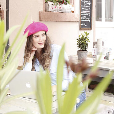 Eine Frau mit Hut sitzt draußen in einem Café, auf ihrem Tisch steht ein Laptop