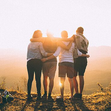 Vier junge Erwachsene stehen Arm in Arm auf einem Hügel und blicken in die Sonne. 