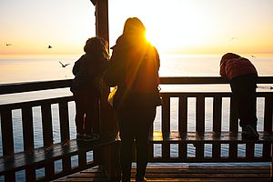 Foto: Unsplash/ Marco Ceschi Eine Frau und zwei Kinder blicken dem Sonnenuntergang entgegen.