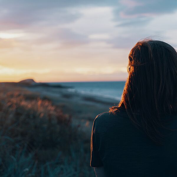 Eine Frau ist bis zum Rücken zu sehen, welche dem Foto abgewandt in Richtung der Sonne blickt, die gerade zwischen Strand und Meer im Hintergrund untergeht.